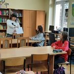 Uczniowie siedzą przy stolikach, nauczycielka stoi trzymając w ręce kartkę. Nauczycielka tłumaczy uczniom zasady konkursu.