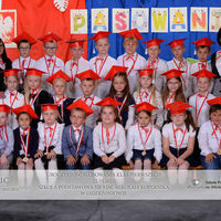 część uczniów klasy 1C siedzi, część stoi, ubrani są na galowo, na szyjach maja medale, na głowach mają czerwone birety, obok stoją ich wychowawczynie, 
