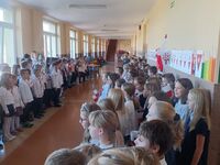 Duża grupa uczniów stoi po dwóch stronach korytarza. Wspólnie śpiewają Hymn Polski.
