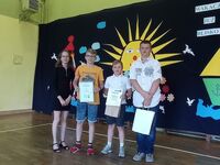 Uczennica i troje uczniów stoją na tle dużego żółtego słońca. Jeden uczeń jest w pomarańczowej koszulce, pozostali uczniowie są w strojach apelowych. Chłopcy trzymają nagrody, które dostali za konkurs.